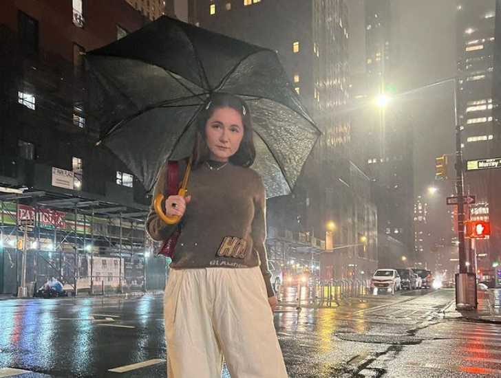 Picture of Emma Rose Kenny holding umbrella in middle of road where it is raining with cars and tall buildings in background